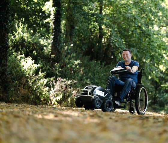 Wheelchair user with wheelchair traction device on a hiking trail in the Southern Eifel Nature Park, © Naturpark Südeifel, Thomas Urbany